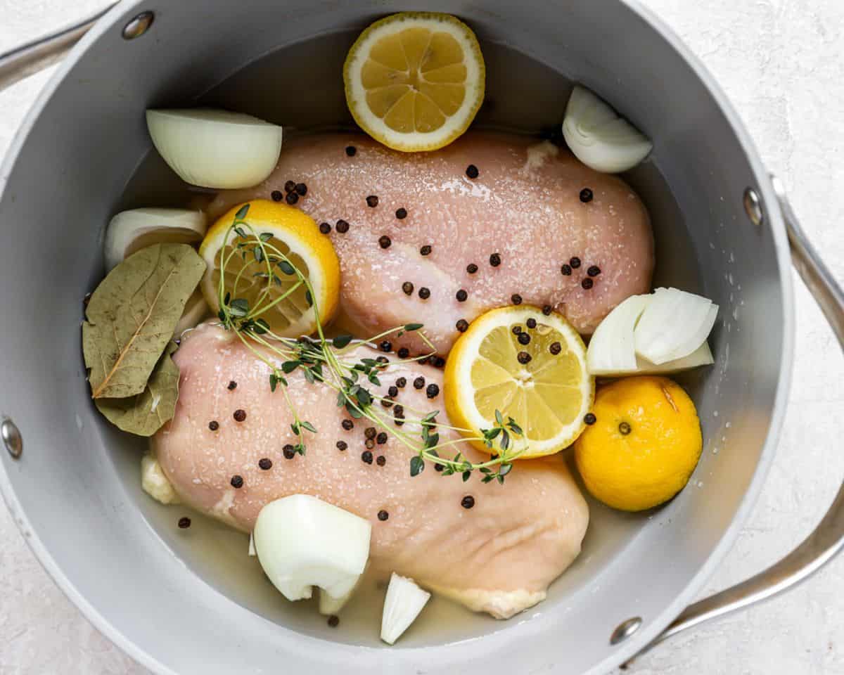 Raw chicken breasts in a pot with lemon slices, onion, bay leaves, peppercorns, and herbs before poaching