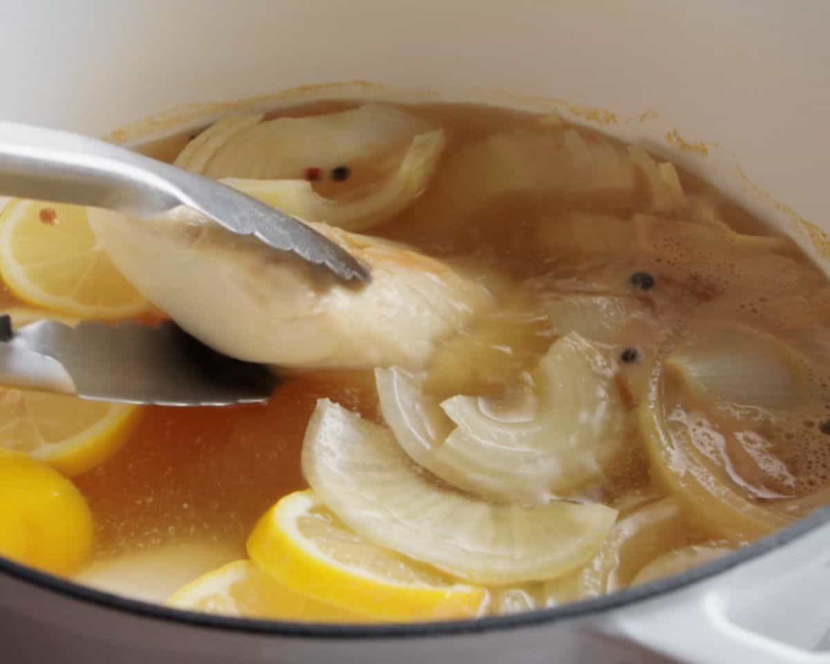Poached chicken breast being lifted from a pot of broth with onions, lemon, and peppercorns