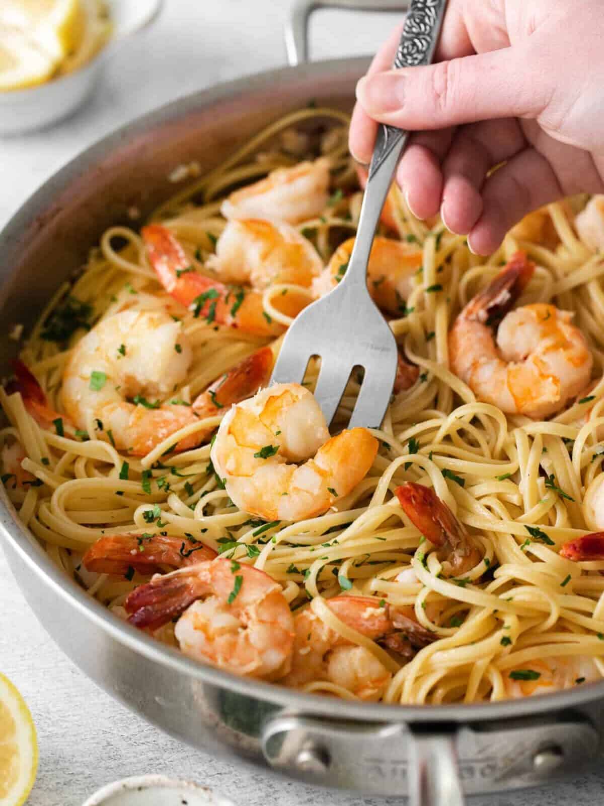 a hand using a slotted spoon to scoop shrimp scampi from a bed of pasta in a frying pan.