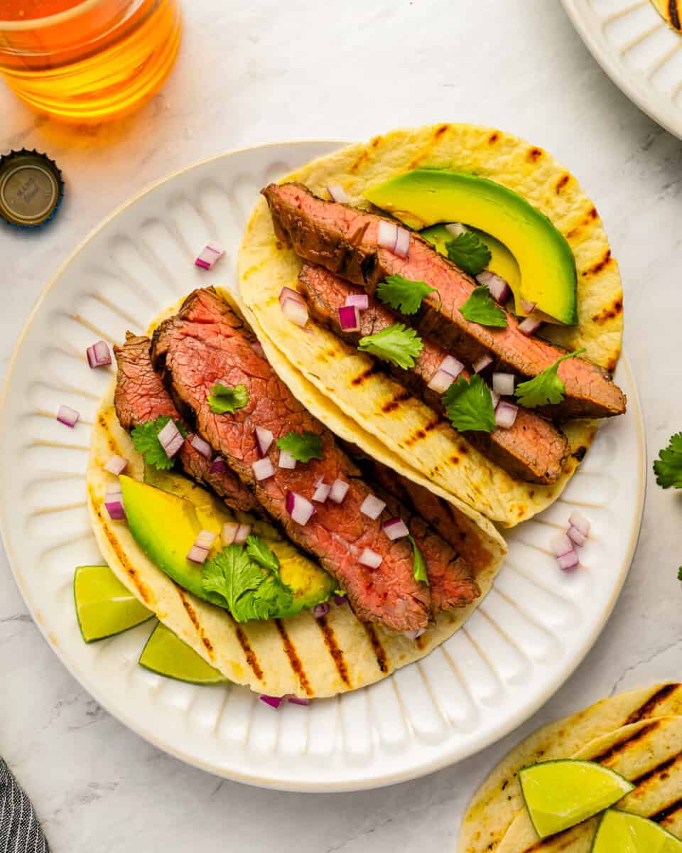 Grilled steak tacos with avocado and lime on a white plate.