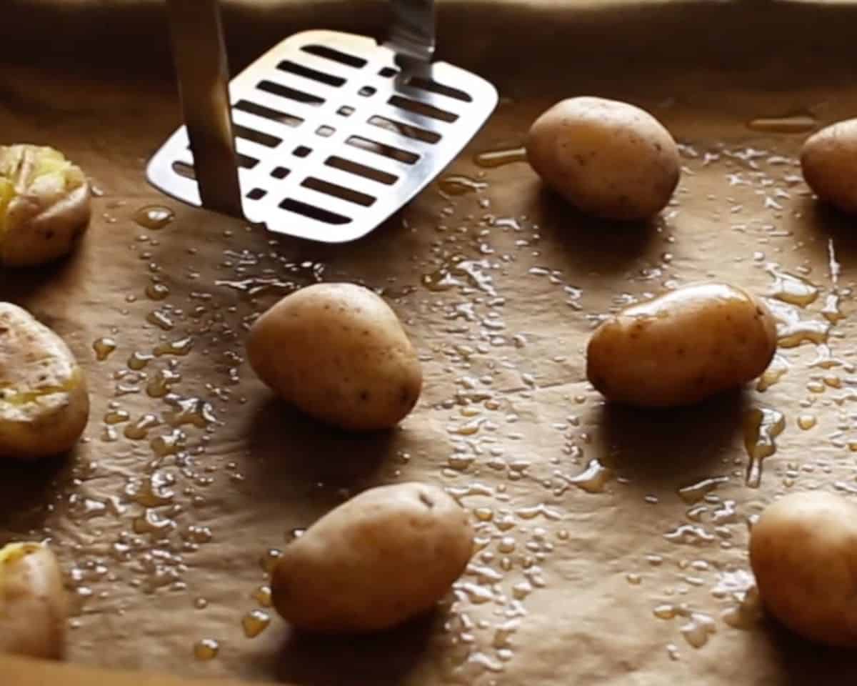 A potato masher pressing down on a boiled baby potato on an oiled parchment sheet pan. - 7