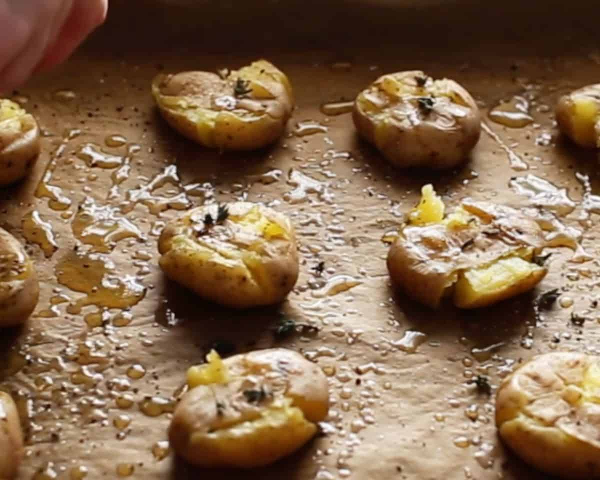 Smashed baby potatoes drizzled with olive oil and sprinkled with herbs on a baking sheet before roasting. - 8
