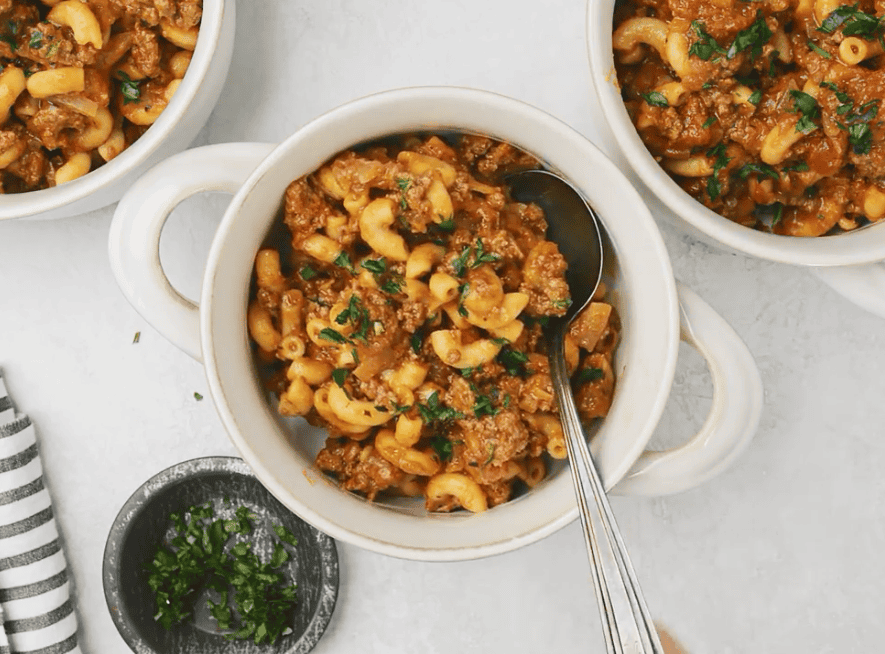 bowls of American Goulash with elbow macaroni, ground beef, and tomato sauce, garnished with fresh parsley, with a spoon in the center bowl and a small dish of chopped parsley alongside.