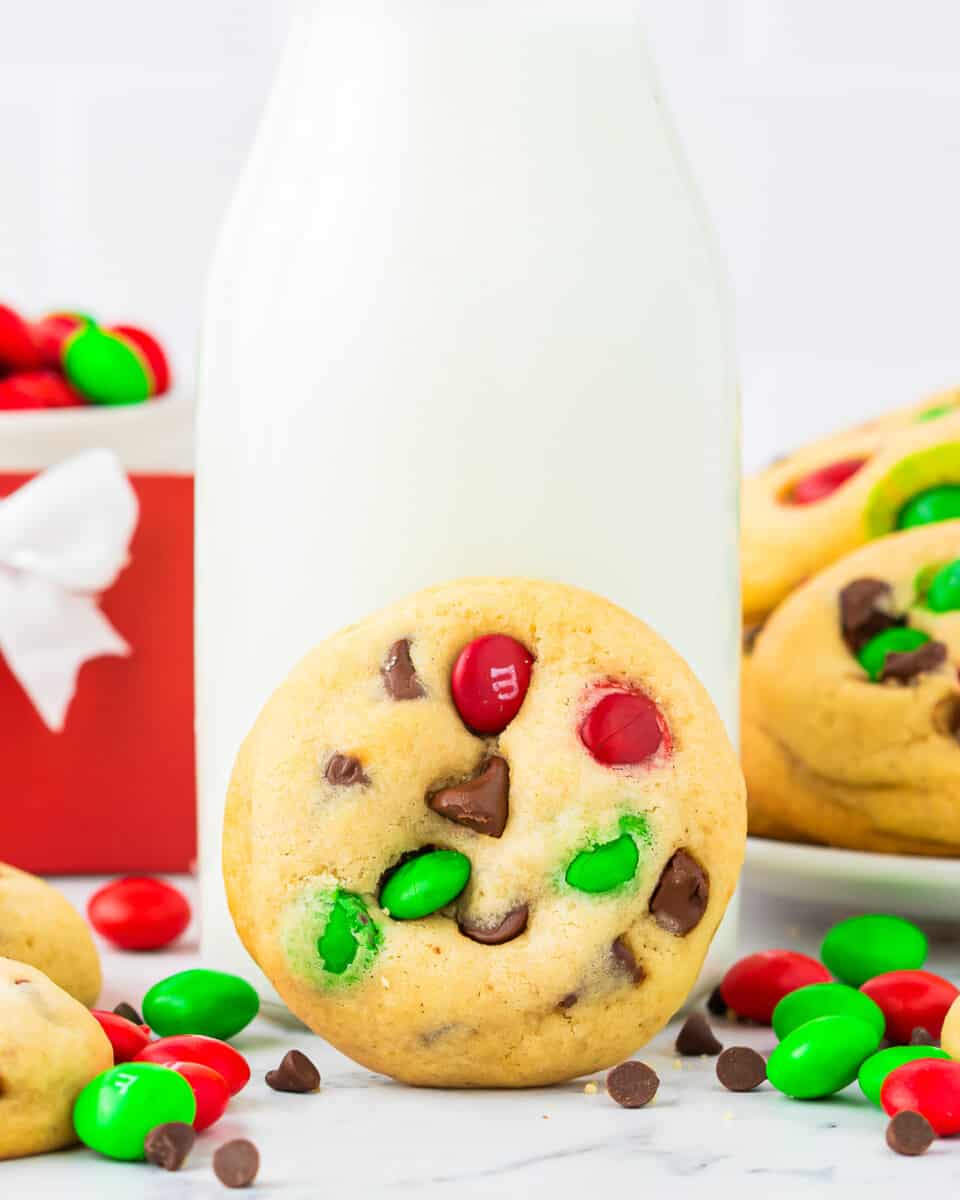 A Christmas M&M cookie leaned up against a jar of milk, with M&Ms and chocolate chips scattered around it.