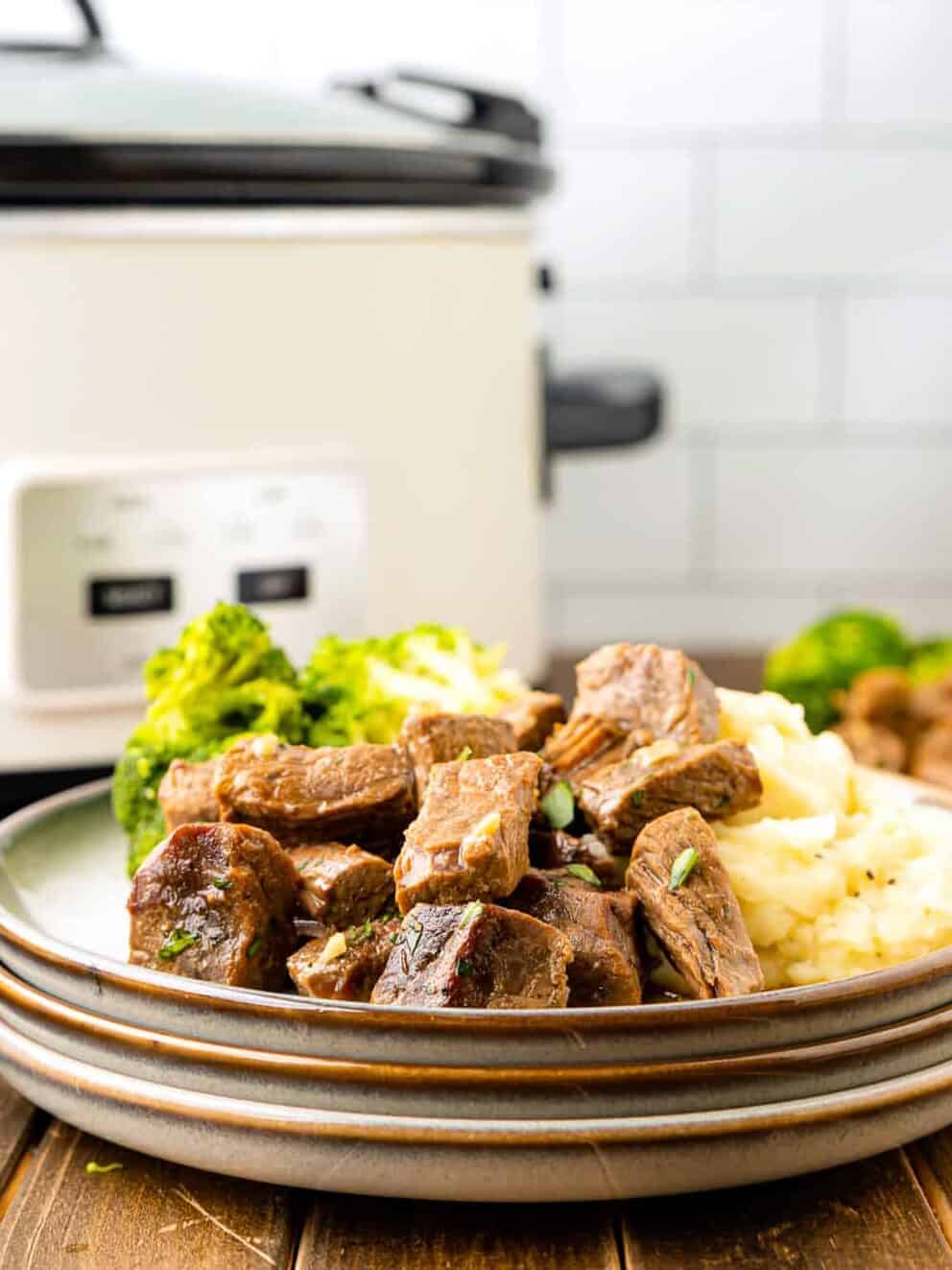 side view of crockpot steak bites on a white plate with mashed potatoes and broccoli.