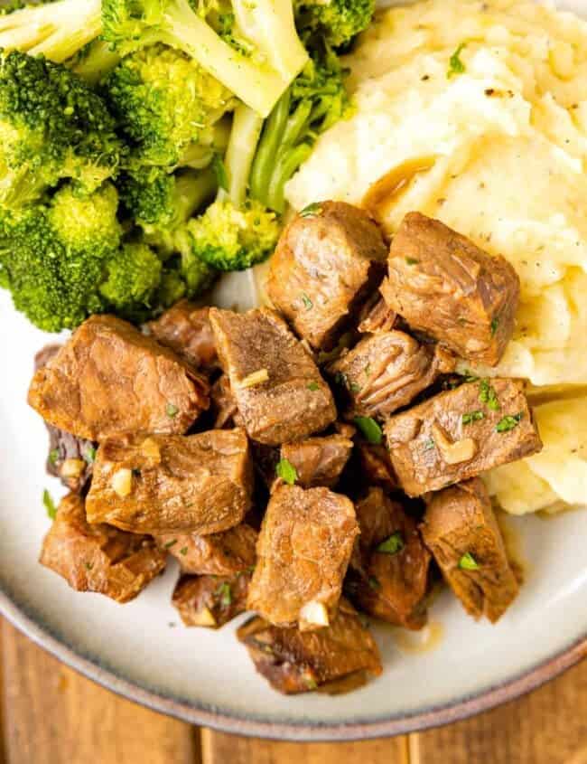 crockpot steak bites on a white plate with mashed potatoes and broccoli.