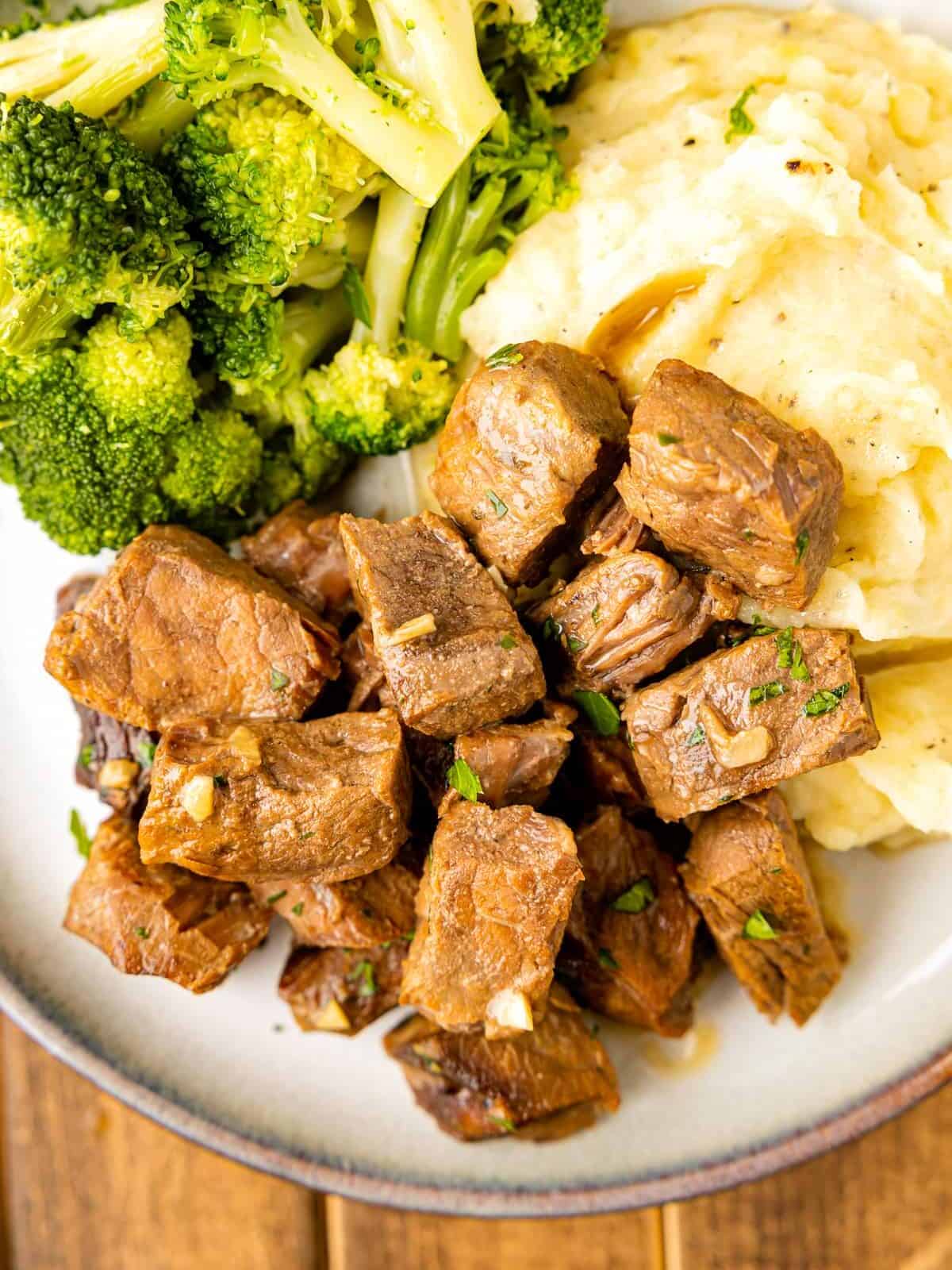 crockpot steak bites on a white plate with mashed potatoes and broccoli.
