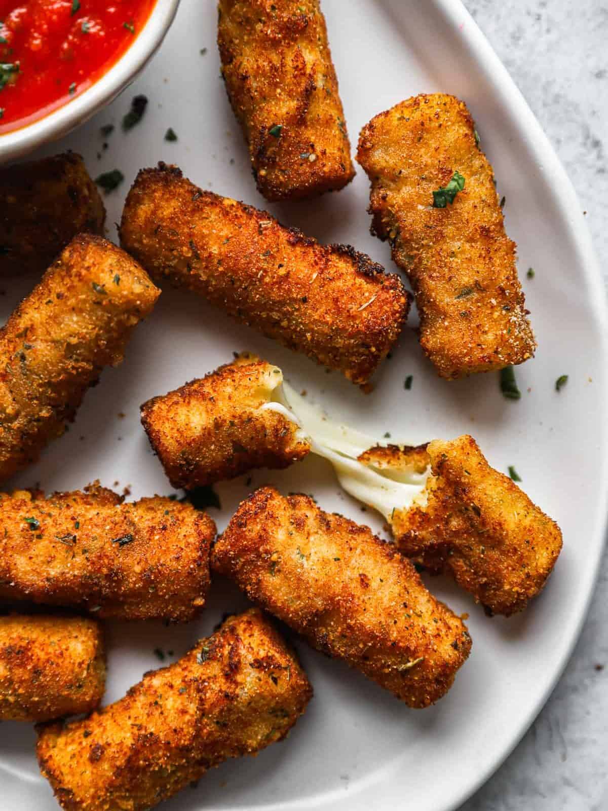 close up overhead view of homemade mozzarella sticks on a white serving platter with a bowl of marinara sauce, one is halved.