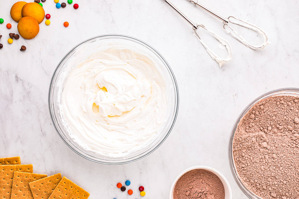 A bowl of whipped cream and cream cheese combined, a bowl of chocolate pudding mix, and a few graham crackers.