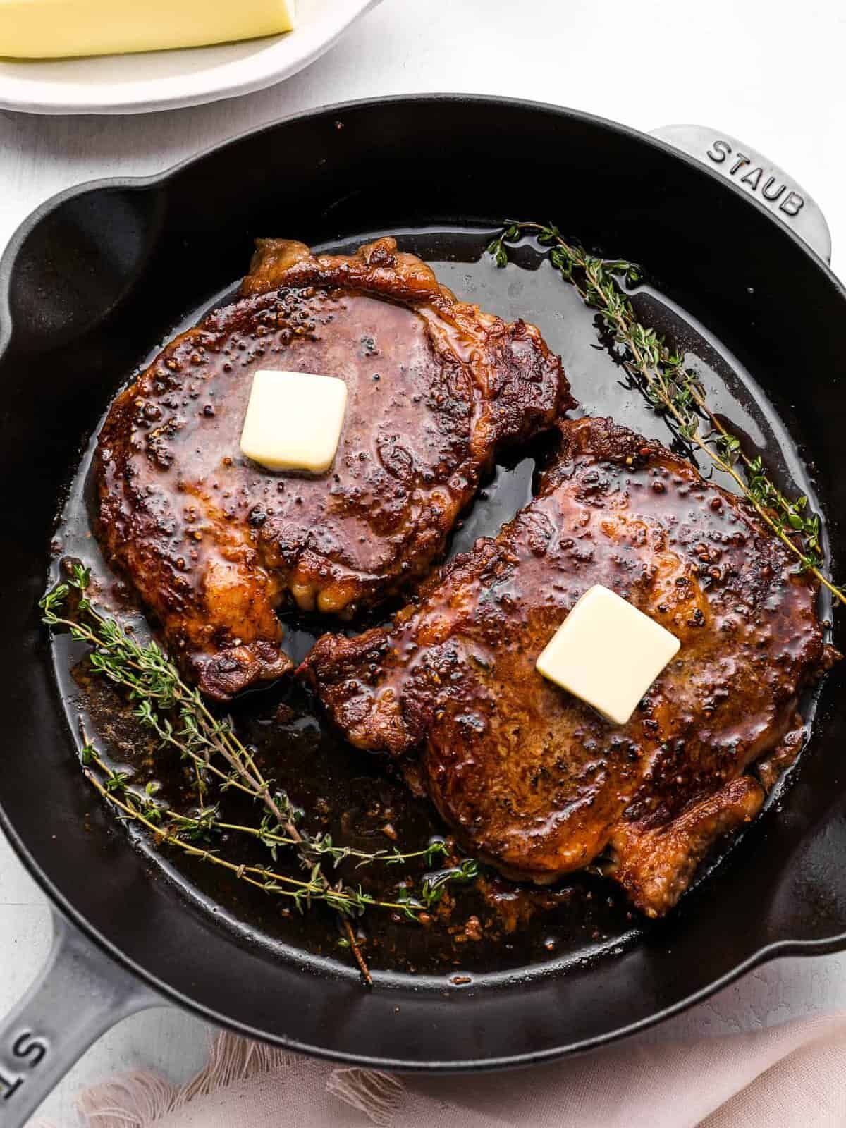 overhead view of oven baked steaks in a cast iron pan topped with a pat of butter and thyme.