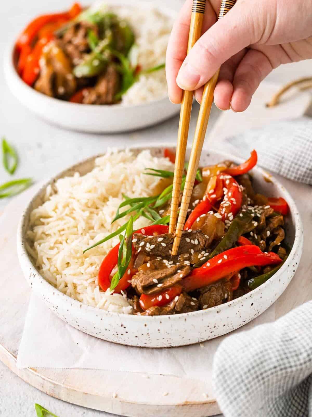 a person holding chopsticks over a bowl of asian stir fry.