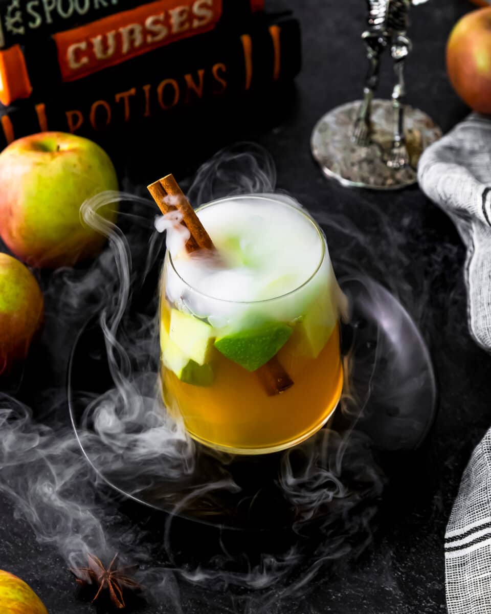 A smoking glass of poison apple sangria with apples and cinnamon on a black background.