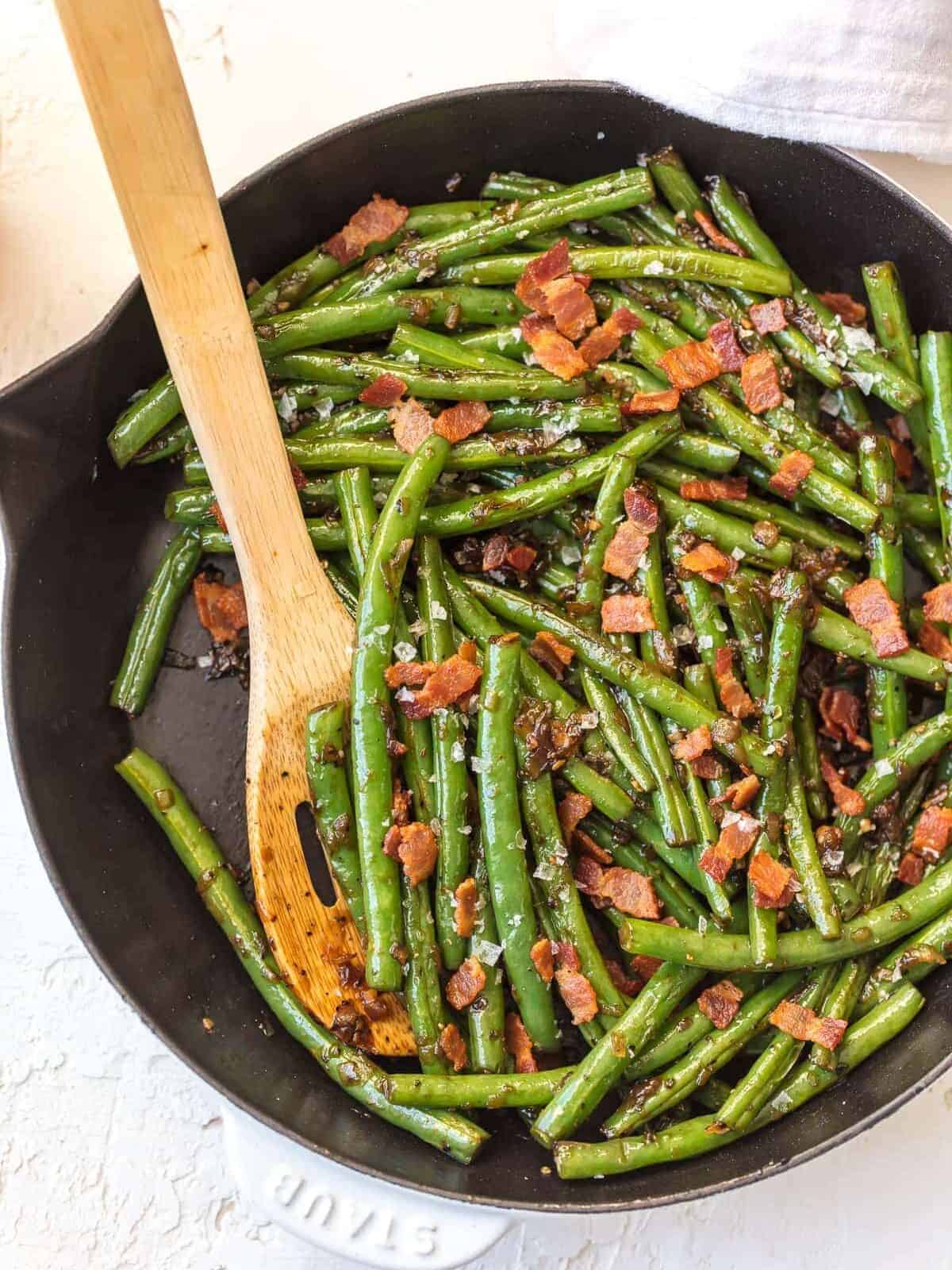 bourbon green beans and bacon in skillet with wooden spoon.