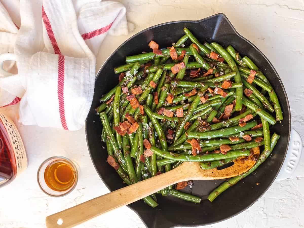 green beans and bacon in a skillet, set on a table top next to a dish towel and a bottle of bourbon.