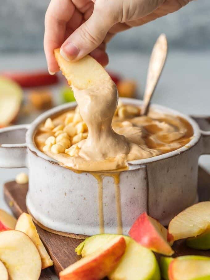 Caramel Apple Dip is a Skinny (believe it!) Caramel Sauce for Apples that will leave your sweet tooth satisfied and is so easy! This Cream Cheese Apple Dip Recipe is what every party needs! This Caramel Apple Dip is a lightened up version of our favorite sweet snack is gone in minutes every time. apple being dipped into caramel apple dip