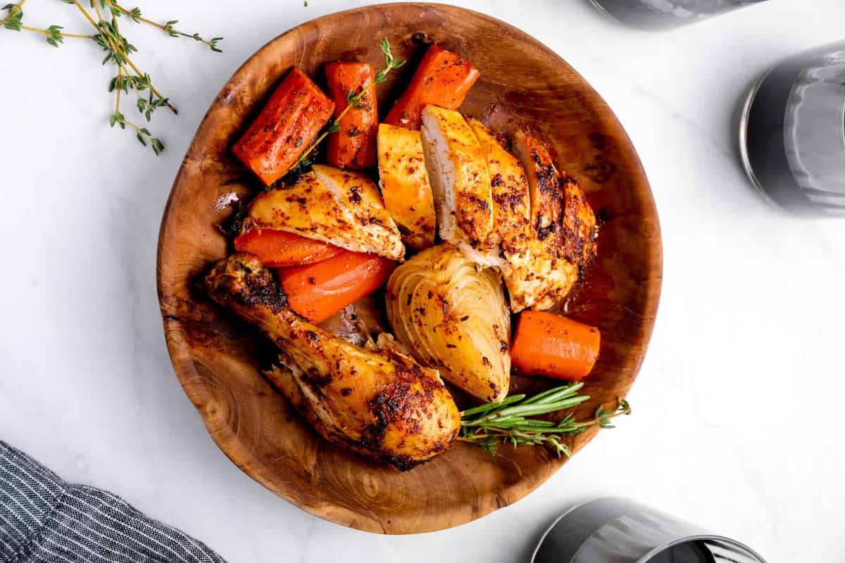 sliced crockpot whole chicken in a wooden bowl with vegetables and herbs.