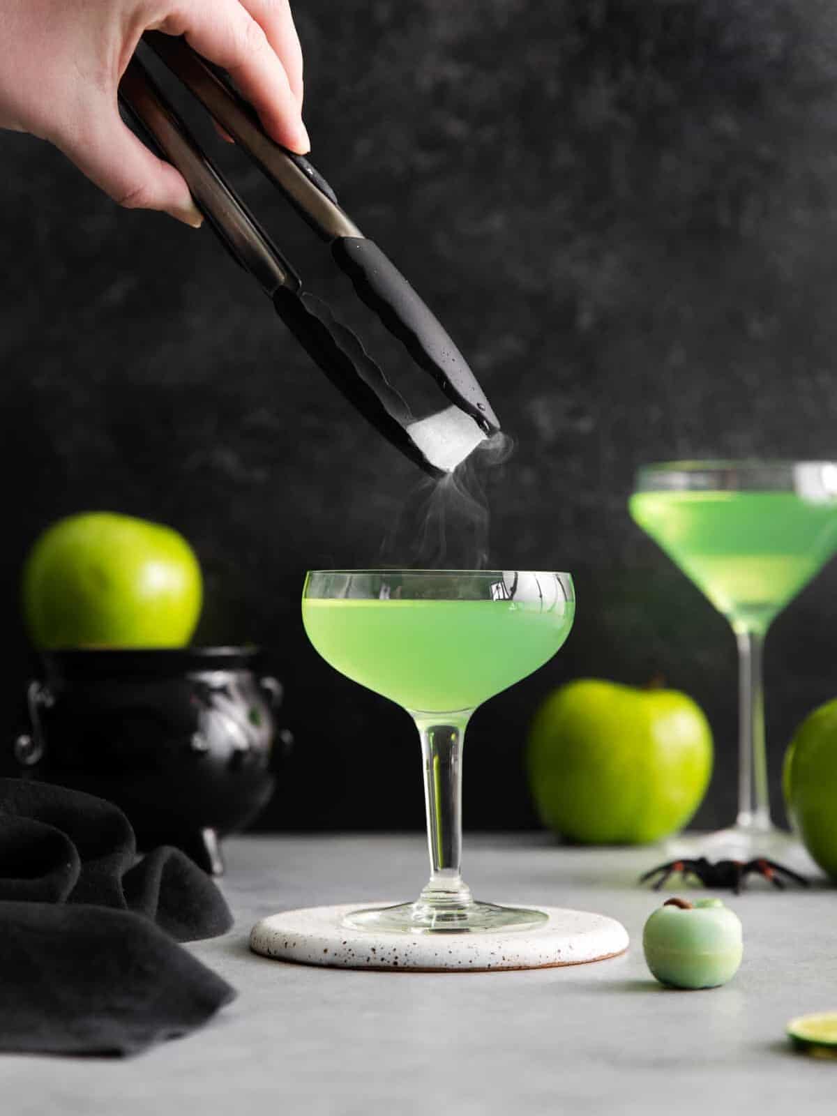 a hand using tongs to drop dry ice into a halloween poison apple martini on a white coaster.