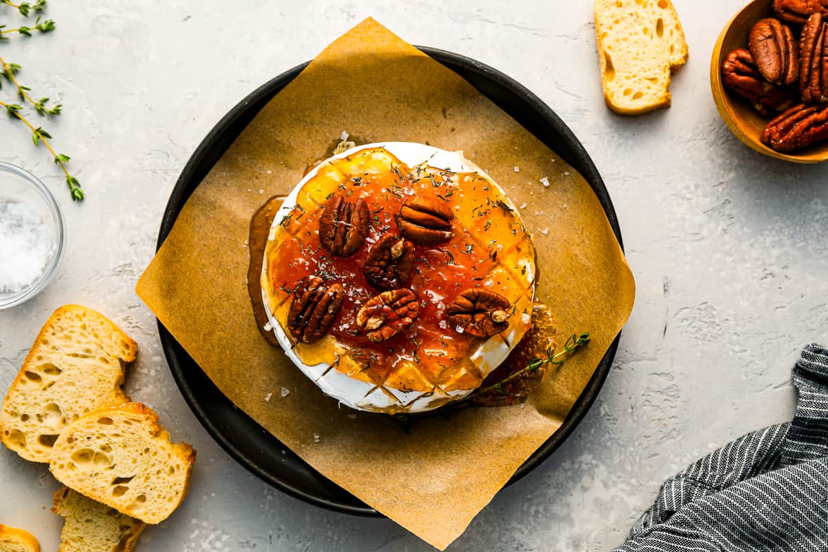 Baked brie at the center of a countertop, surrounded by slices of bread, thyme, and pecans.