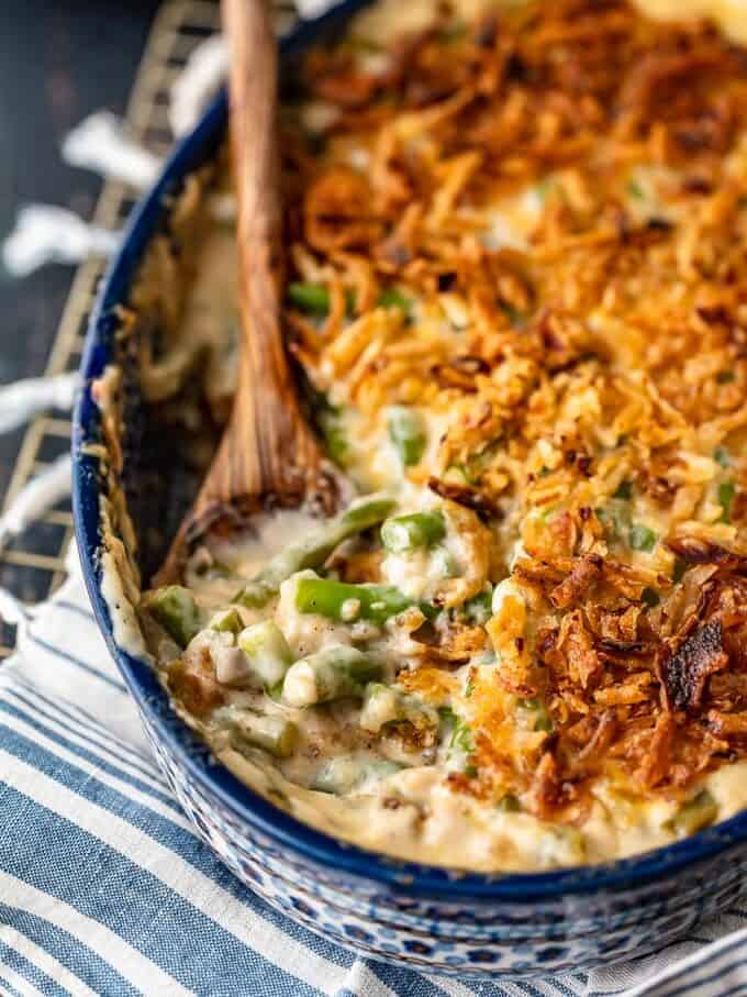 Classic Green Bean Casserole is one of those Thanksgiving recipes that you don't need to reinvent. It's absolutely perfect just the way it is, with green beans, cream of mushroom, crunchy fried onions, and some other awesome ingredients. This green bean casserole recipe is exactly what you need for you next holiday meal! green bean casserole in a blue dish with a wooden spoon.