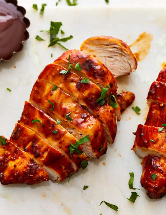 Two air fryer chicken breasts on a cutting board with BBQ sauce and herbs.