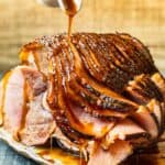 hand pouring glaze onto honey baked ham on a white serving platter