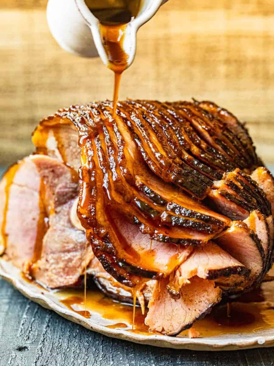 hand pouring glaze onto honey baked ham on a white serving platter