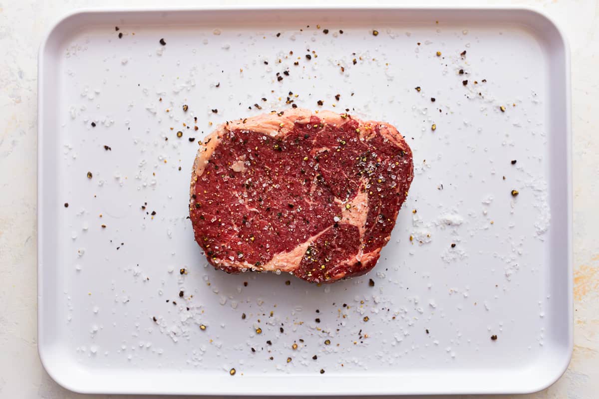 A piece of seasoned ribeye steak on a white plate.