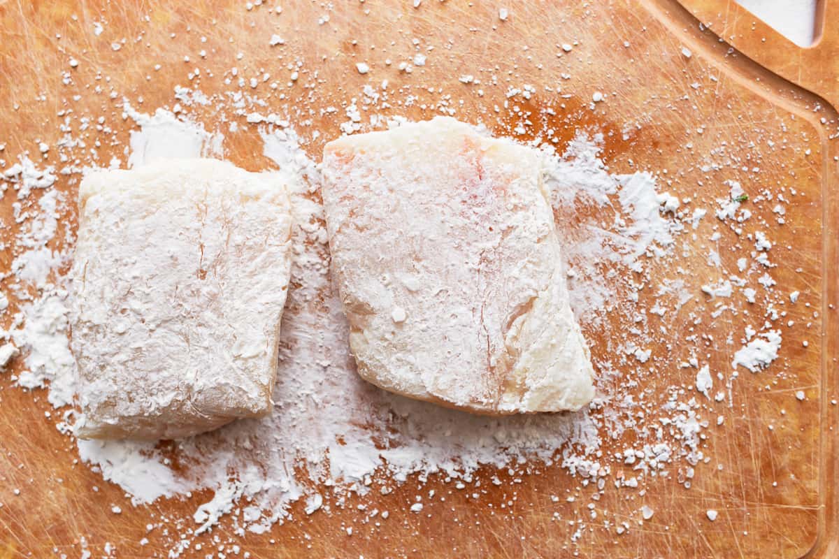 Two pieces of fish on a wooden cutting board, coated in cornstarch.