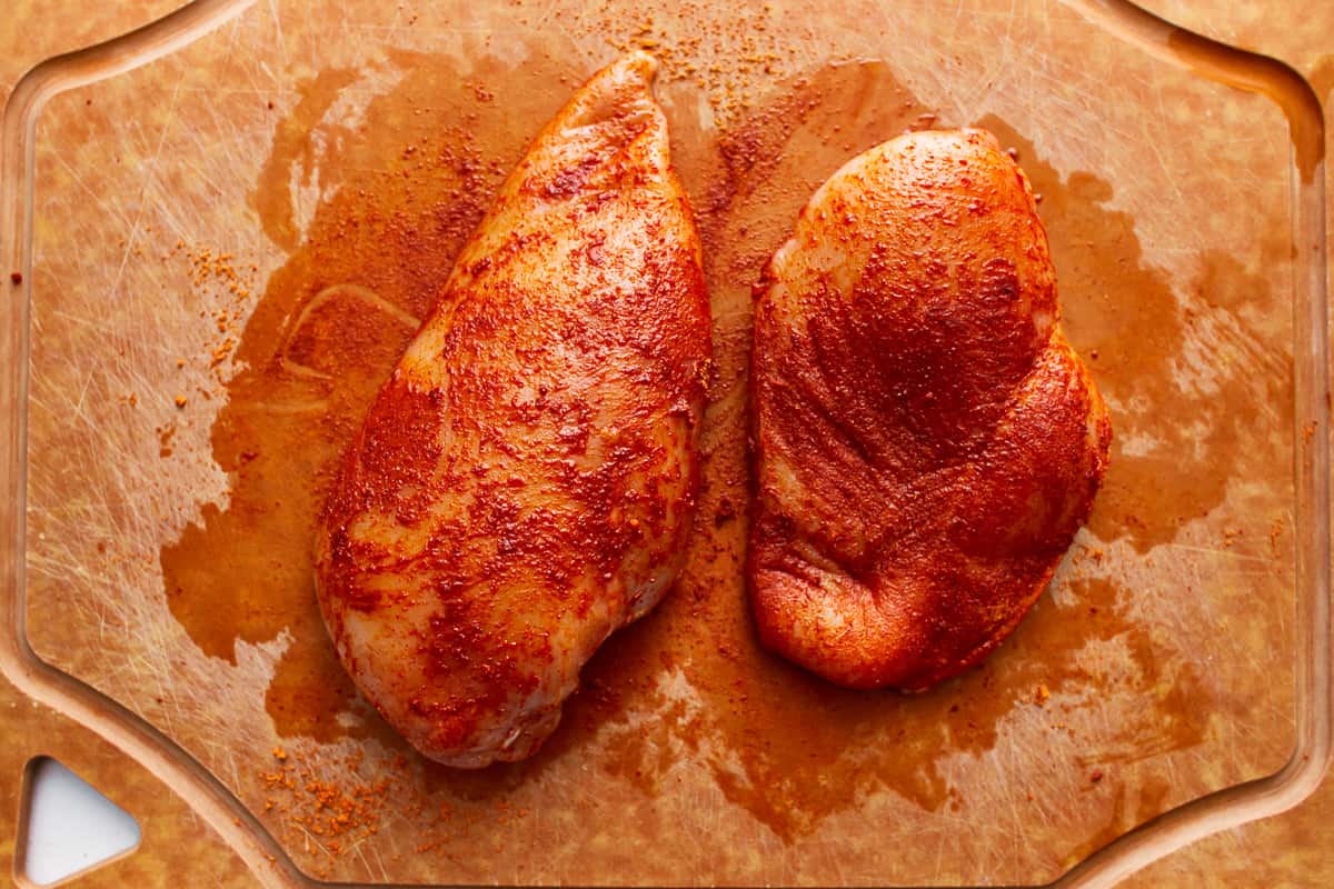 Two seasoned chicken breasts on a wooden cutting board.