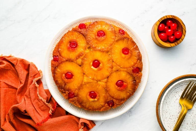 A whole pineapple upside-down cake on a white plate, topped with circular pineapple slices and maraschino cherries in the center of each slice. The cake is accompanied by a small bowl of cherries, a plate, and a set of gold-colored utensils on a white marble surface.