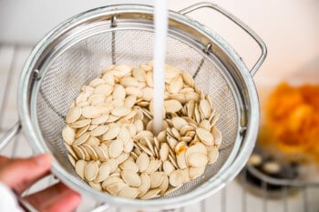 A person pouring liquid into a strainer full of pumpkin seeds.