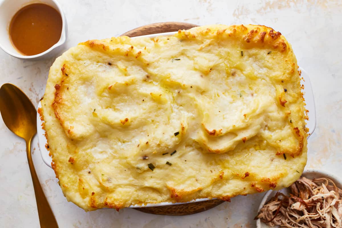 A dish of baked mashed potatoes on a table top, next to dishes of turkey and gravy.