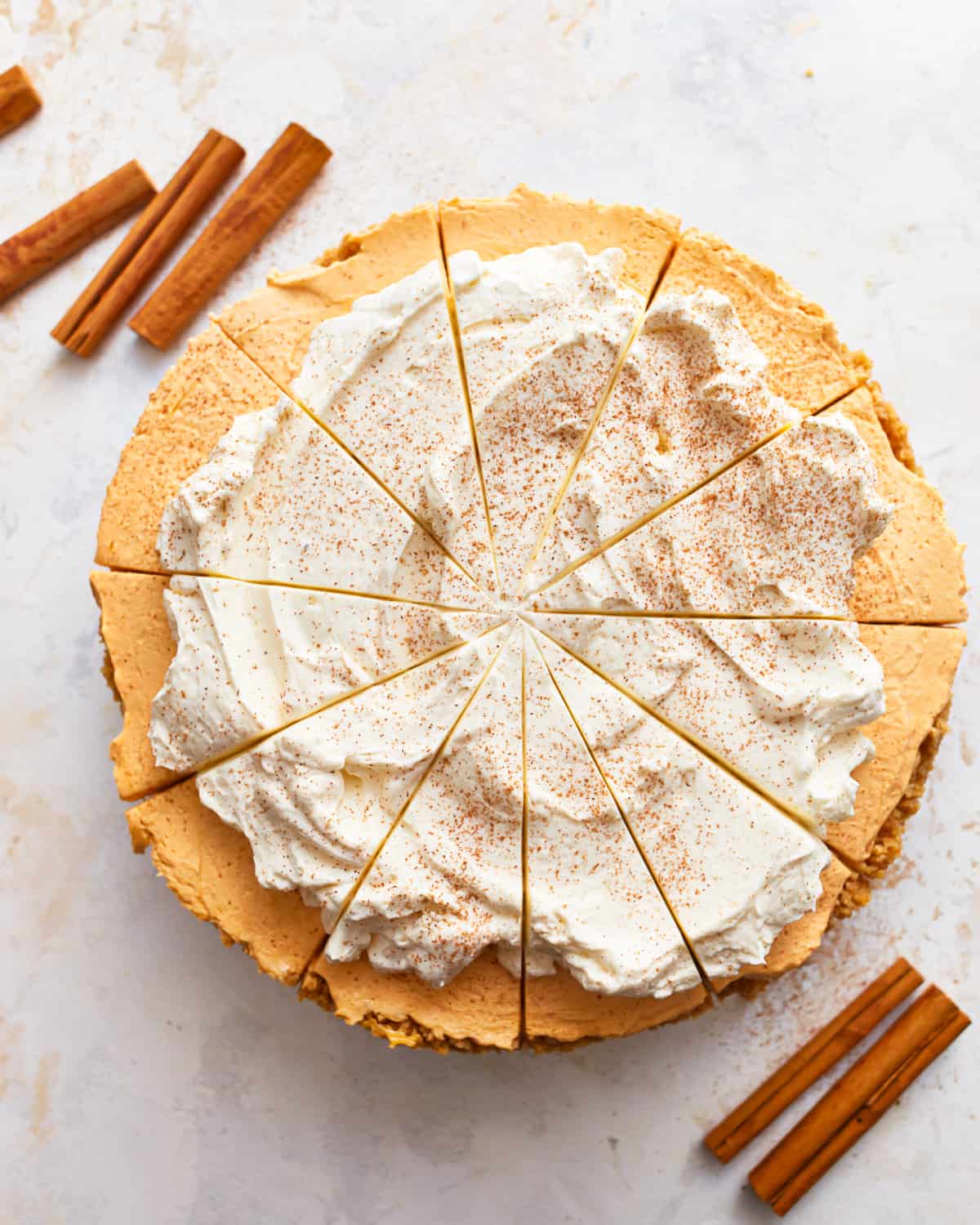 A whole no bake pumpkin cheesecake, cut into slices, topped with whipped topping, and surrounded by cinnamon sticks.