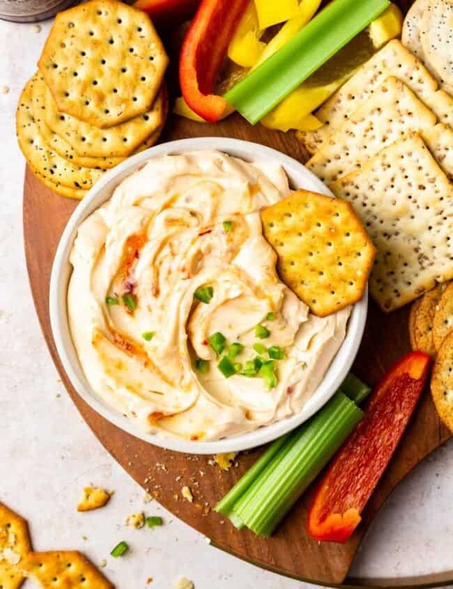 a board of crackers and veggies, with a bowl of pepper jelly cream cheese dip