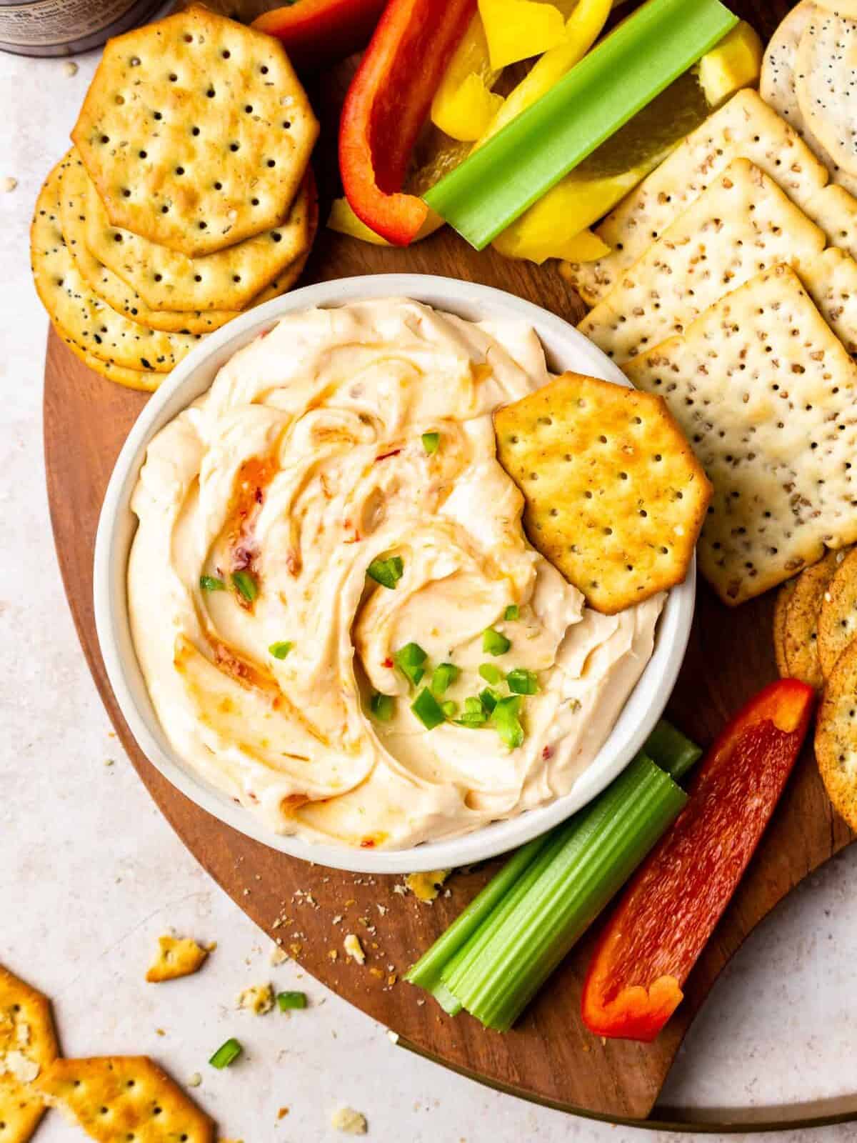 a board of crackers and veggies, with a bowl of pepper jelly cream cheese dip
