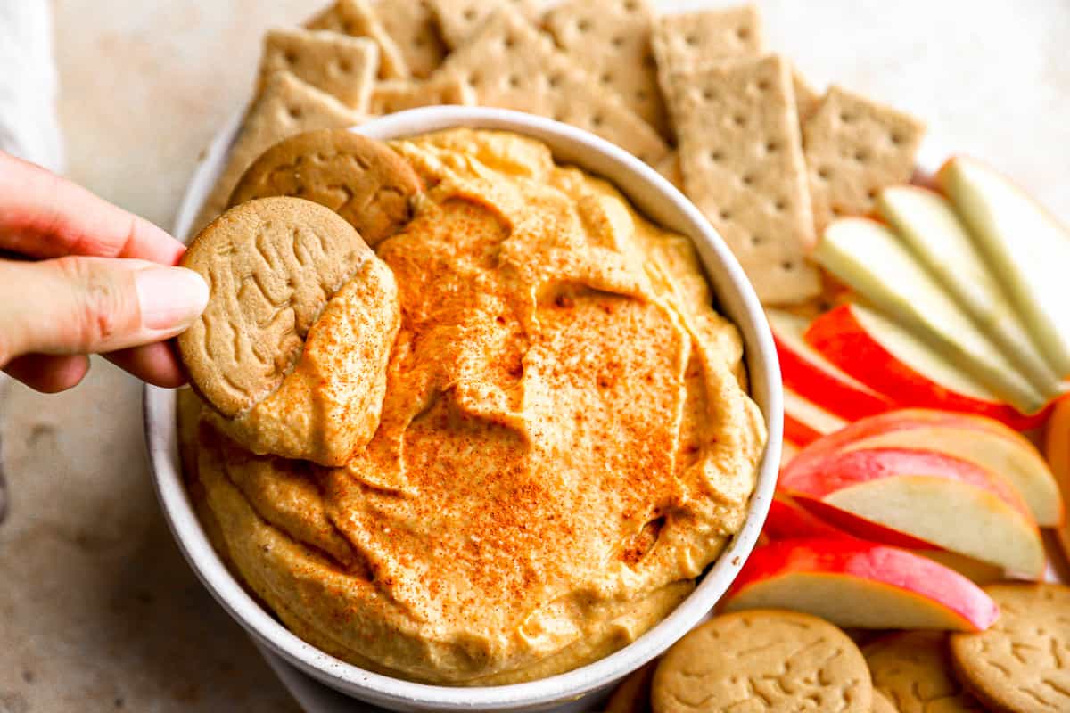 A person dipping a cracker into a bowl of pumpkin cream cheese dip.