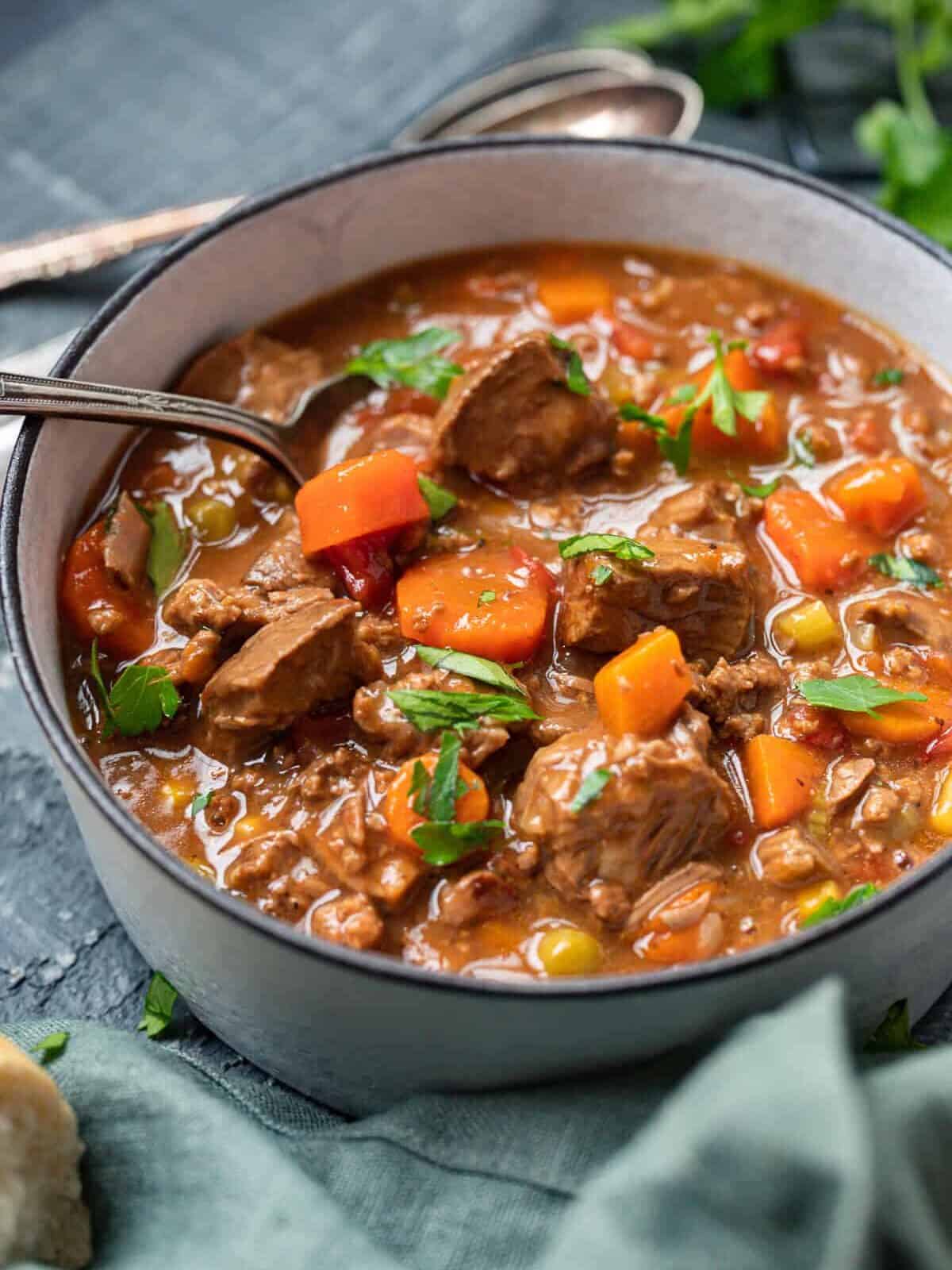 three-quarters view of steak soup in a dutch oven with a serving spoon. Chunks of beef, carrots, and vegetables are visible in the broth.