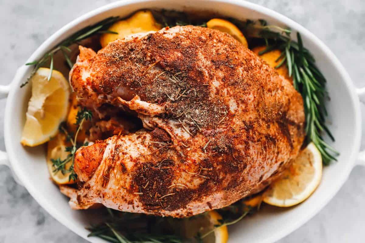 overhead view of a seasoned turkey in a white casserole dish with herbs and lemon wedges.