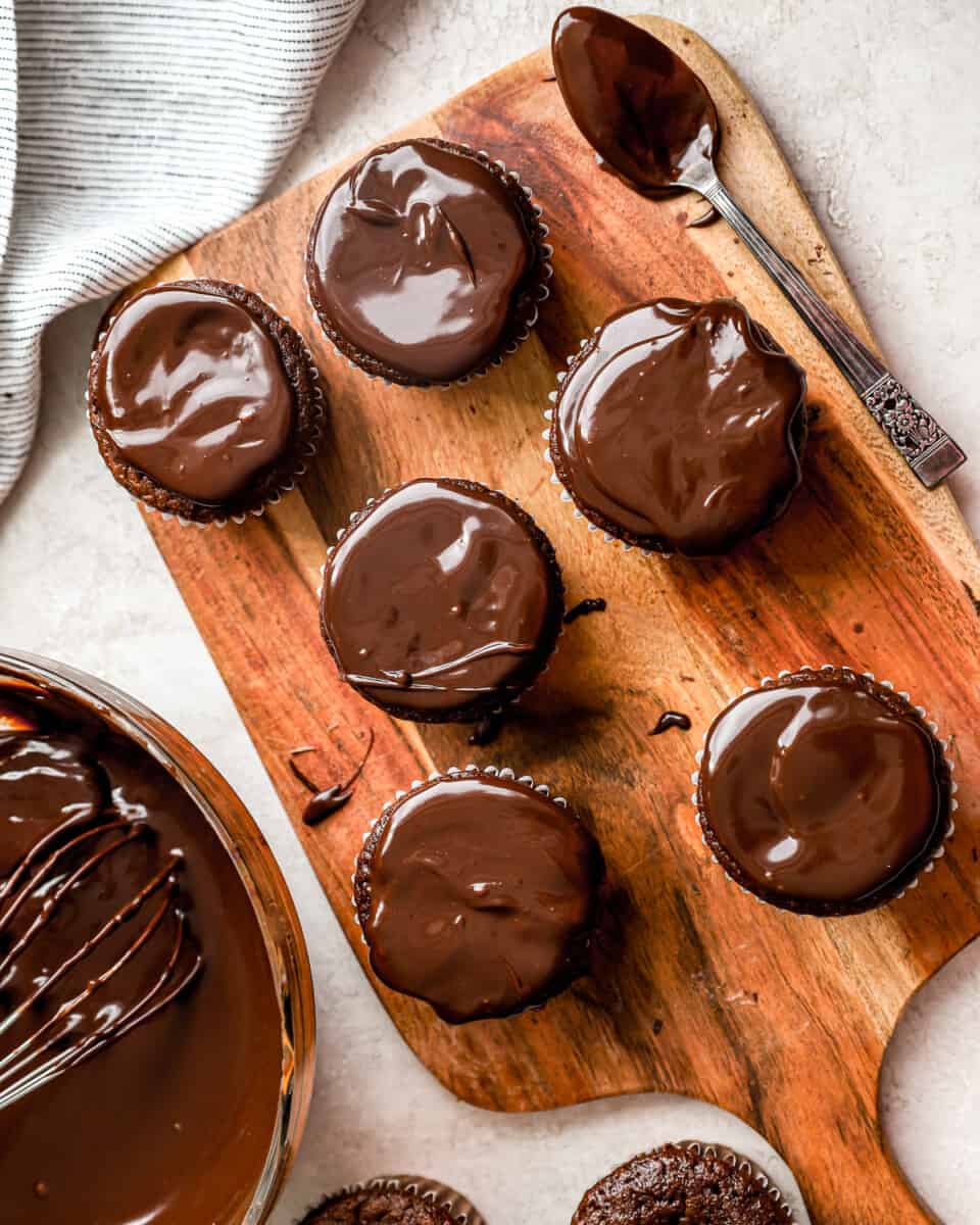 Cupcakes topped with chocolate ganache on a cutting board with a spoon.