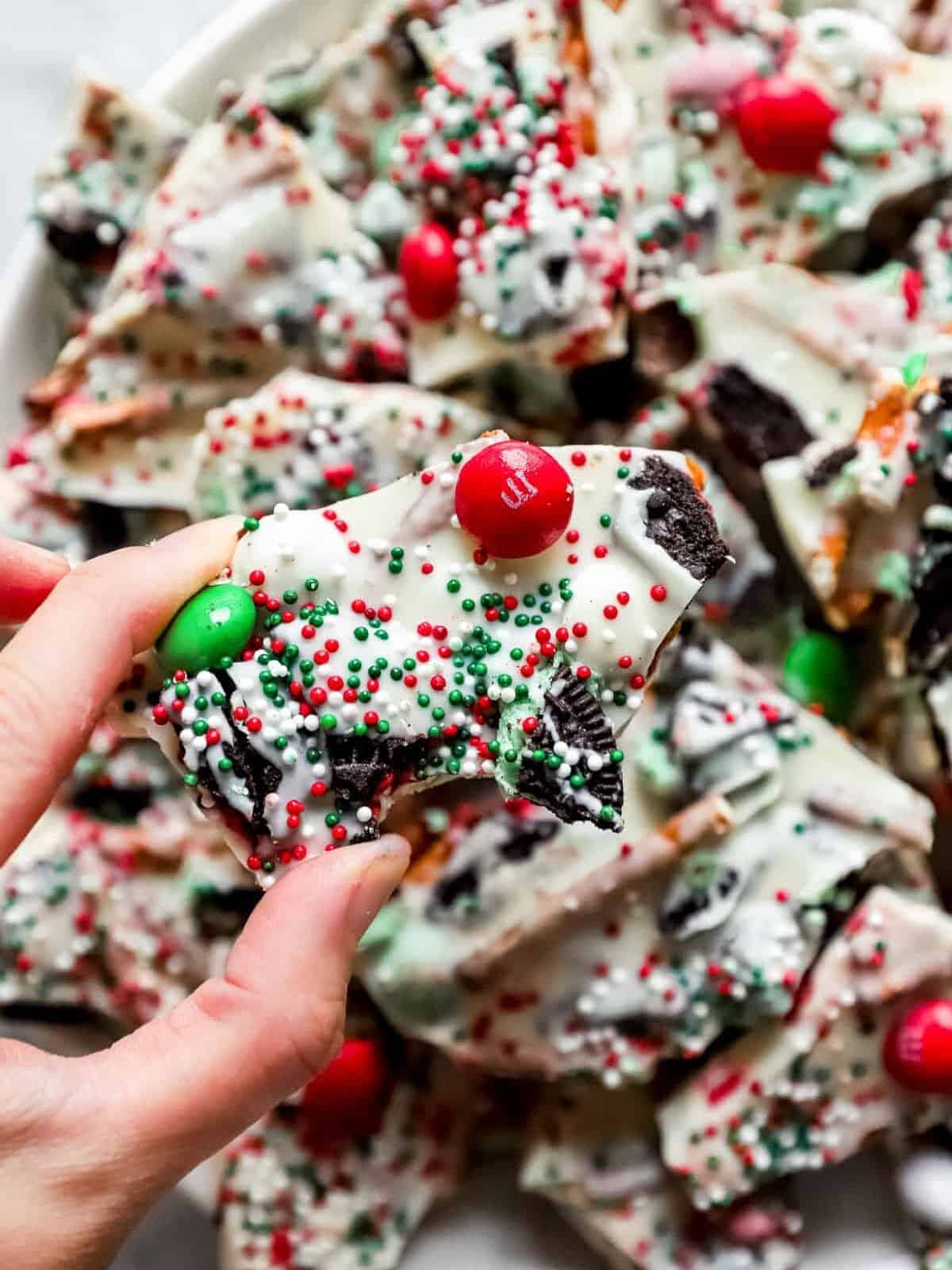 hand holding up a piece of Christmas bark, with M&Ms, sprinkles, and pieces of Oreos