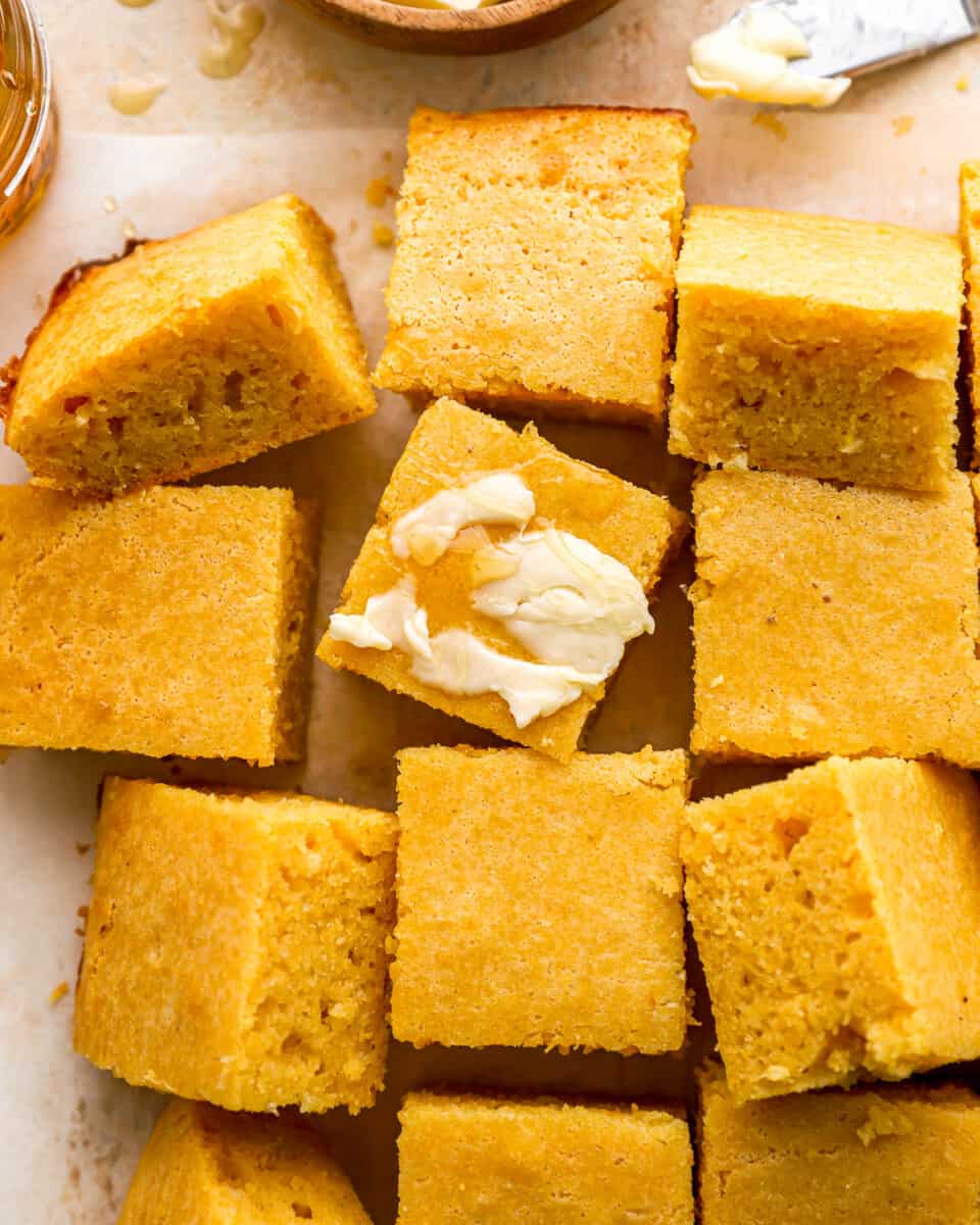 Homemade cornbread squares with butter and honey on a baking sheet.