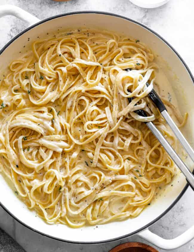 overhead view of fettuccini alfredo in a white dutch oven with tongs.