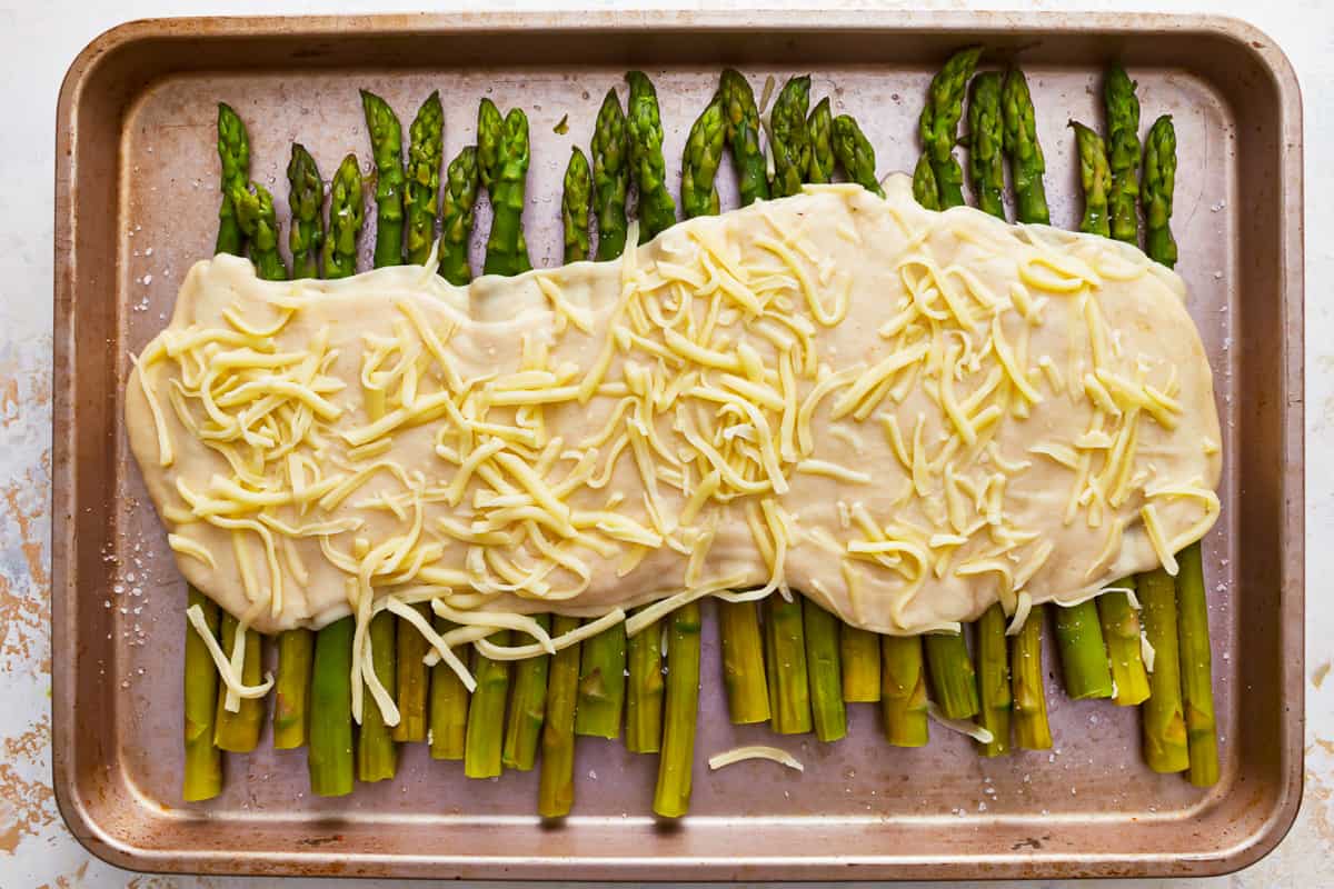 Asparagus topped with cheese sauce on a baking sheet.