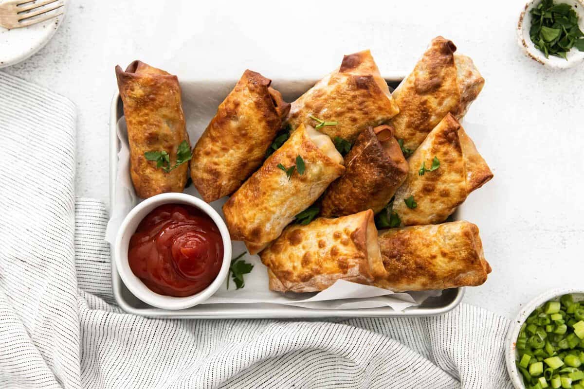 overhead image of egg rolls on a serving tray with a side of ketchup in a white bowl.
