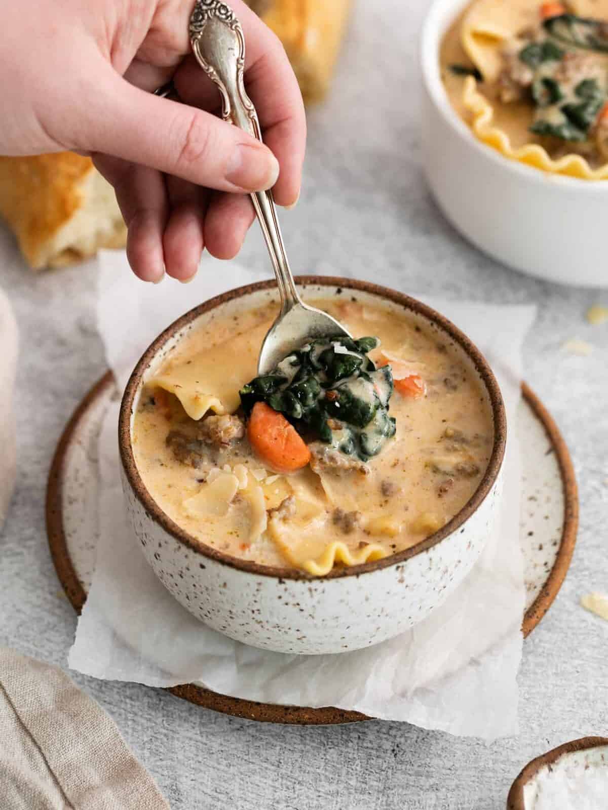 a hand using a spoon to scoop white lasagna soup from a white and brown bowl on a plate.