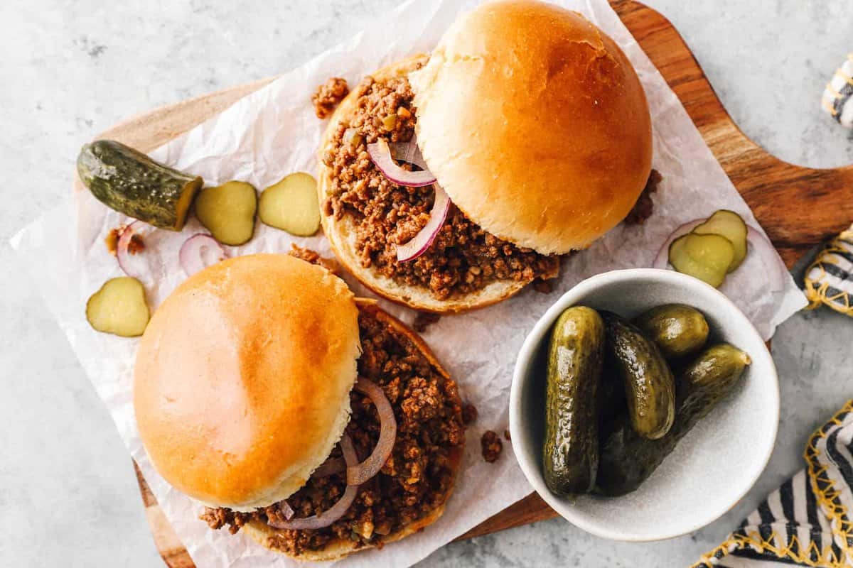 overhead view of 2 sloppy joe sandwiches on a wooden cutting board with pickles.