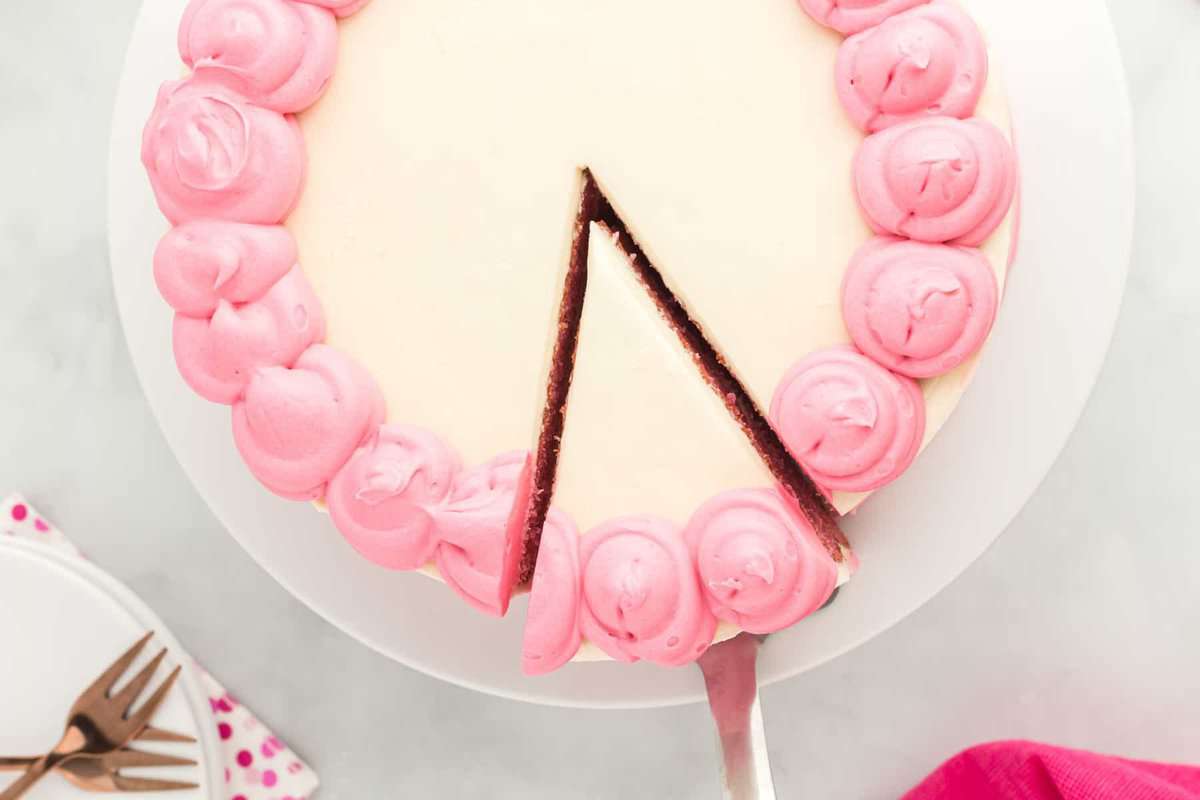 overhead view of a cake server lifting a slice of pink velvet cake from a white cake stand.