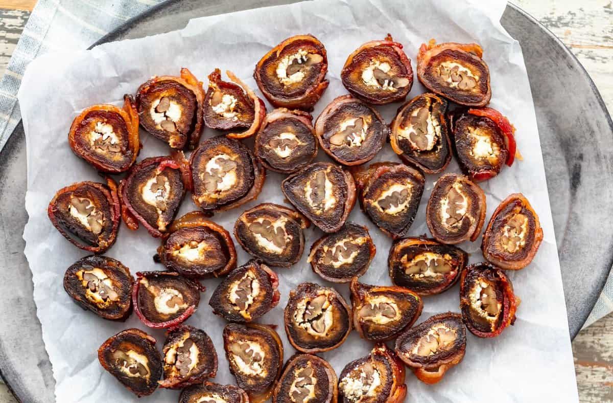 A tray of bacon-wrapped dates is placed on parchment paper. Each date is sliced in half, revealing a visible walnut inside. The presentation is arranged in a slightly scattered pattern.