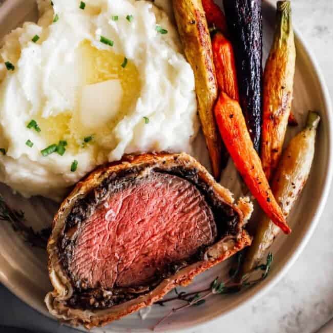 a slice of beef wellington on a white plate with carrots and mashed potatoes.