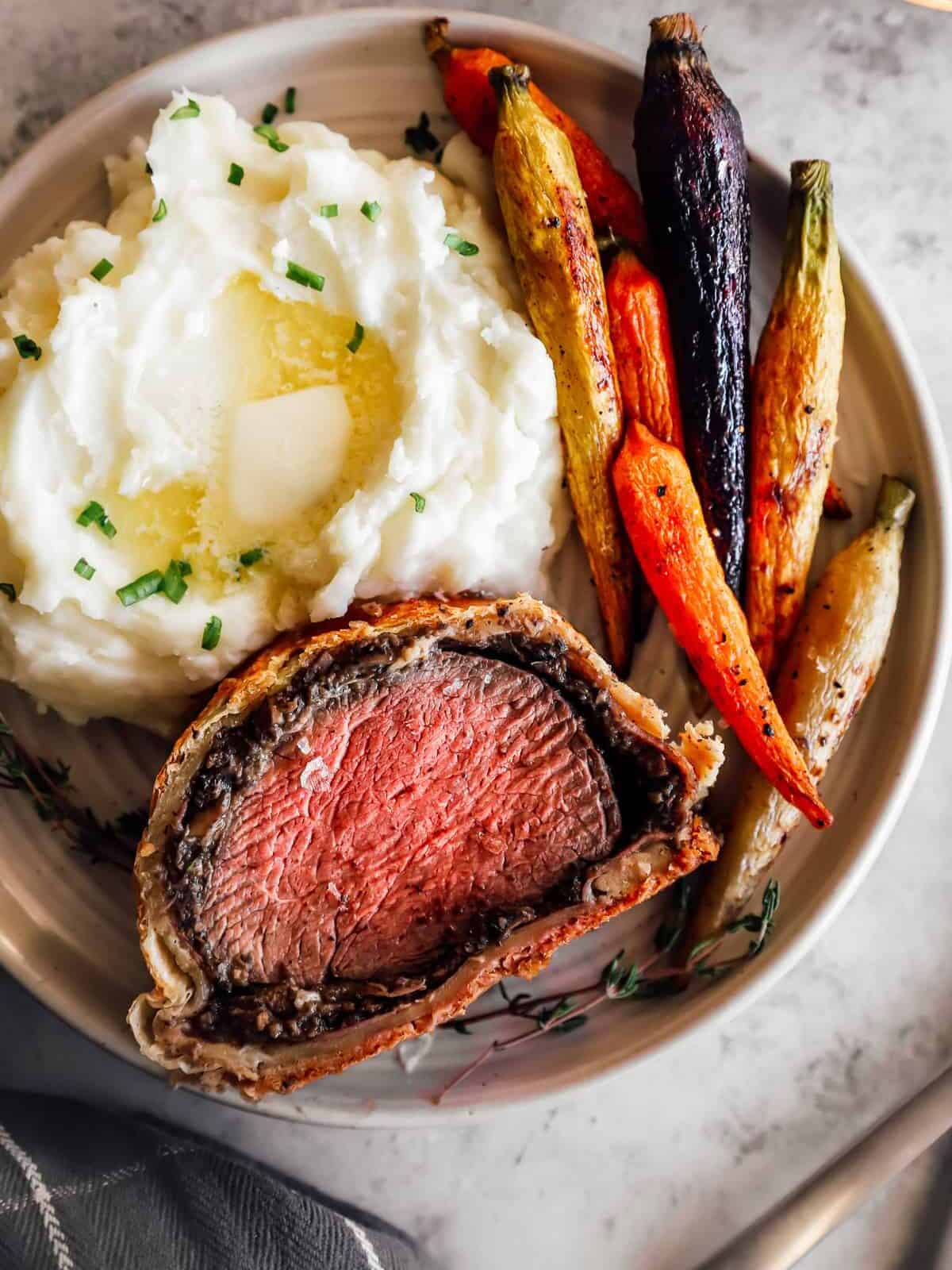 a slice of beef wellington on a white plate with carrots and mashed potatoes.